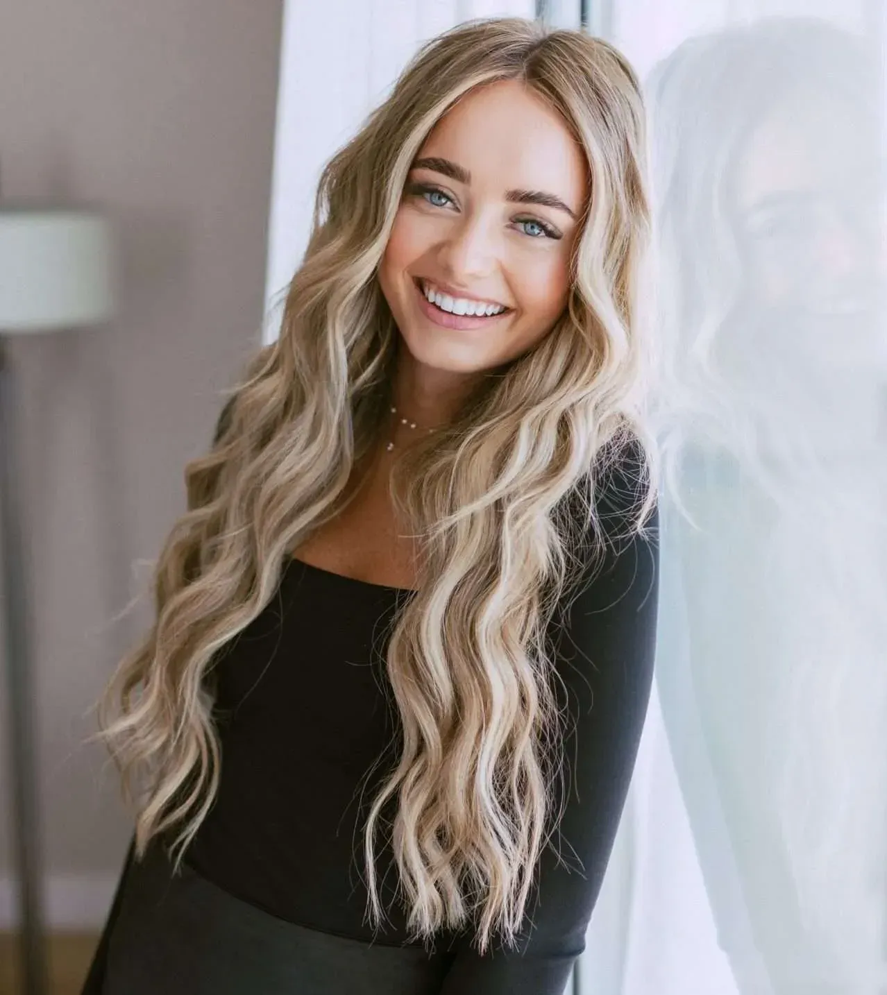 A woman with long blonde hair is smiling and leaning against a window.